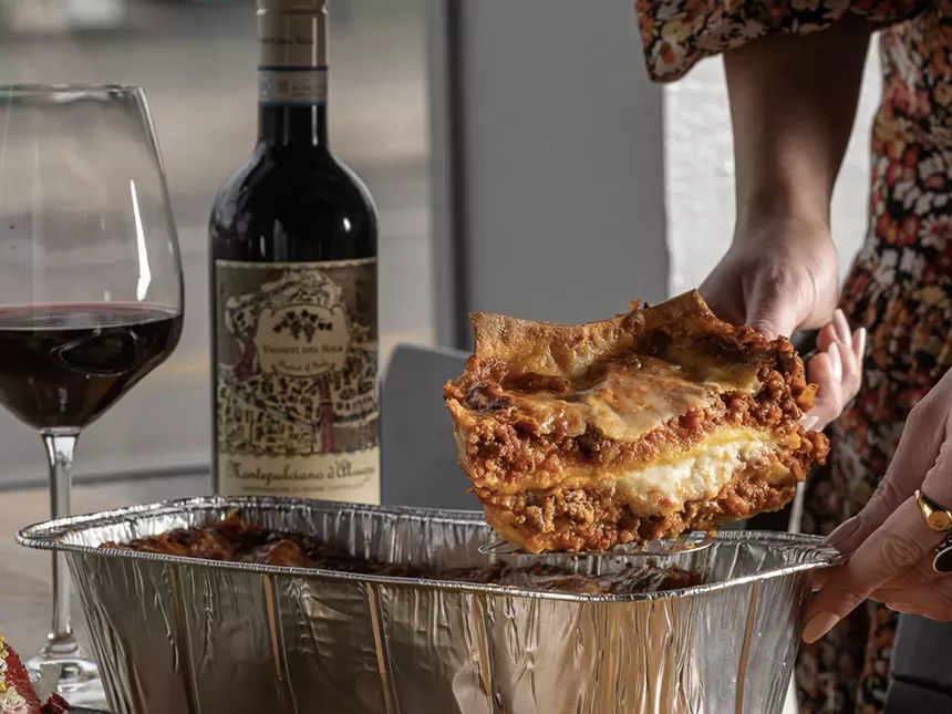 A woman serving a slice of Restaurant Olivia lasagna with a bottle of wine in the background