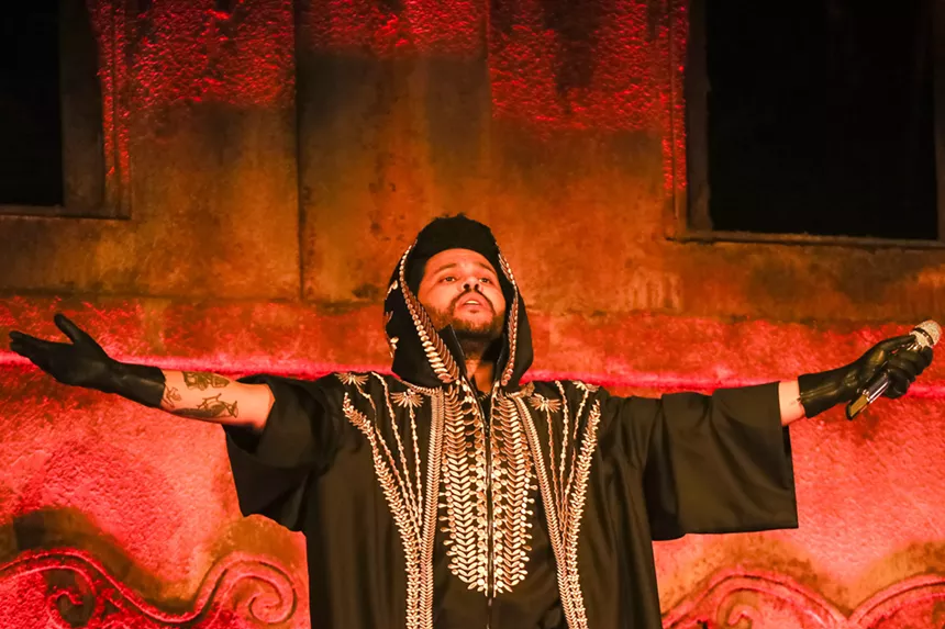 The Weeknd in front of an orange screen with his arms spread open