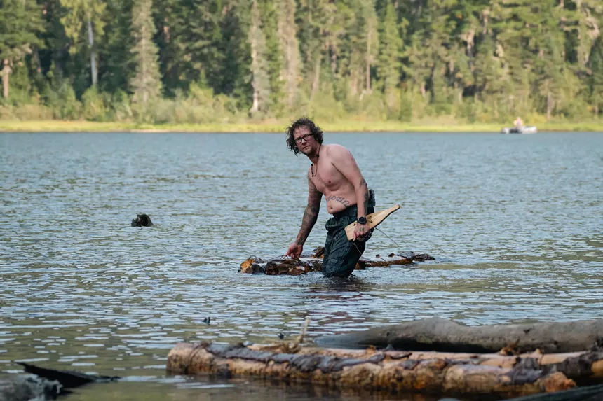 Ryan Willis, shirtless and standing knee-high in the lake with his primitive raft