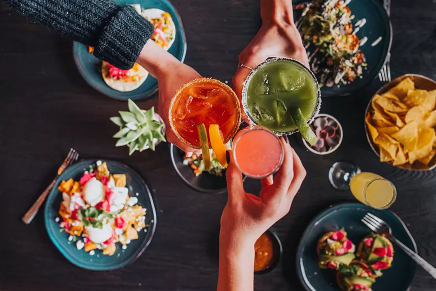 A group of three toasting colorful cocktails above Mexican brunch plates at Tamayo