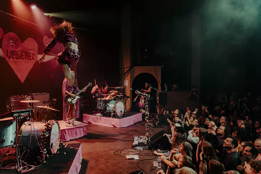 A photo of the Velveteers performing on stage at the Bluebird Theatre, showing the drummer jumping from a platform on stage.