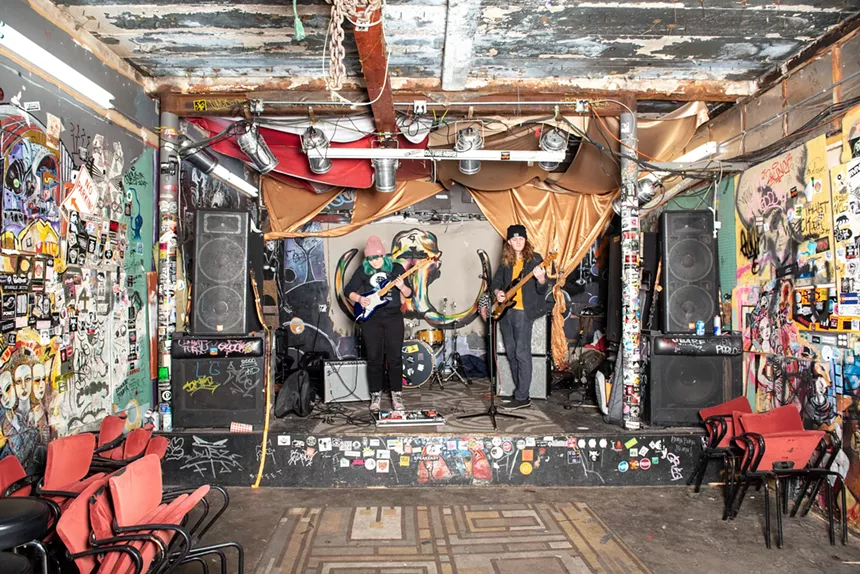 Two musicians playing on a stage, with stickers lining the walls