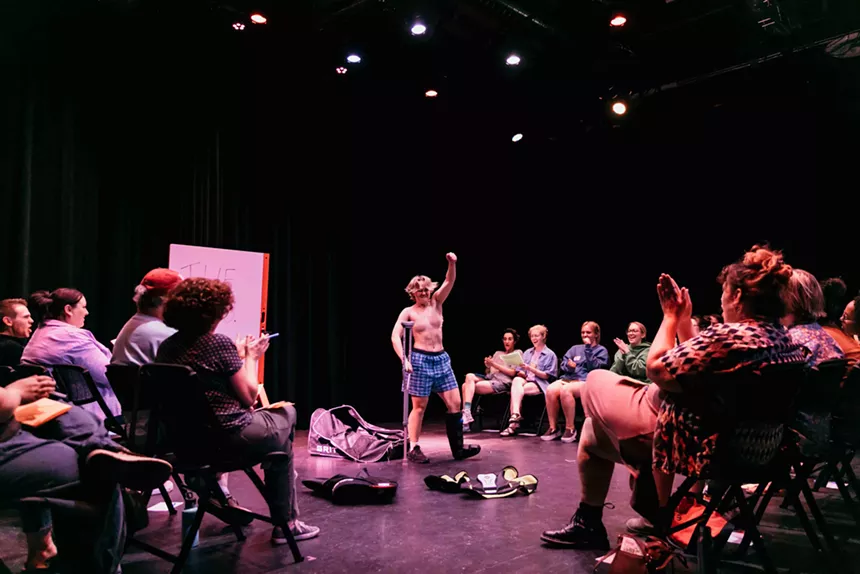 A performer stands onstage, surrounded by a seated audience.