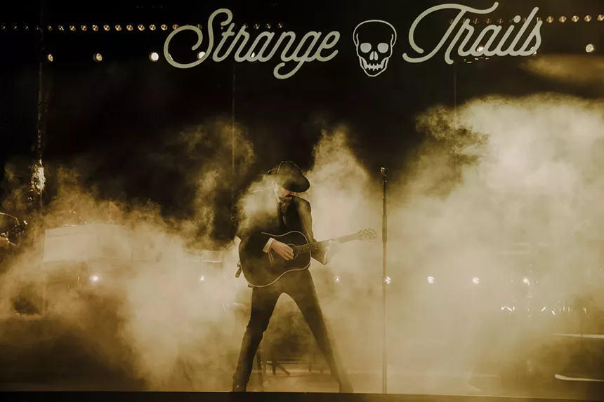 Lord Huron performing on stage at Red Rocks with fog behind him, and a stage sign labeled Strange Trails hangs above him.