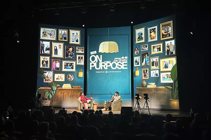 Jay Shetty and Lilly Singh backdropped by an office-like set on three digital panels at the Ellie Caulkins Opera House