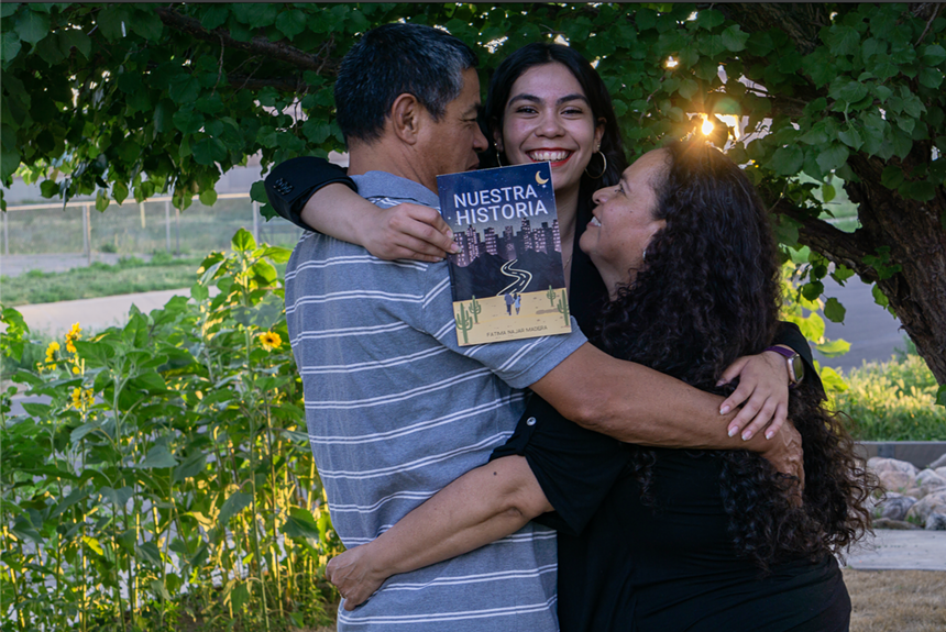 A family of three hug each other under a tree while a book is being held.