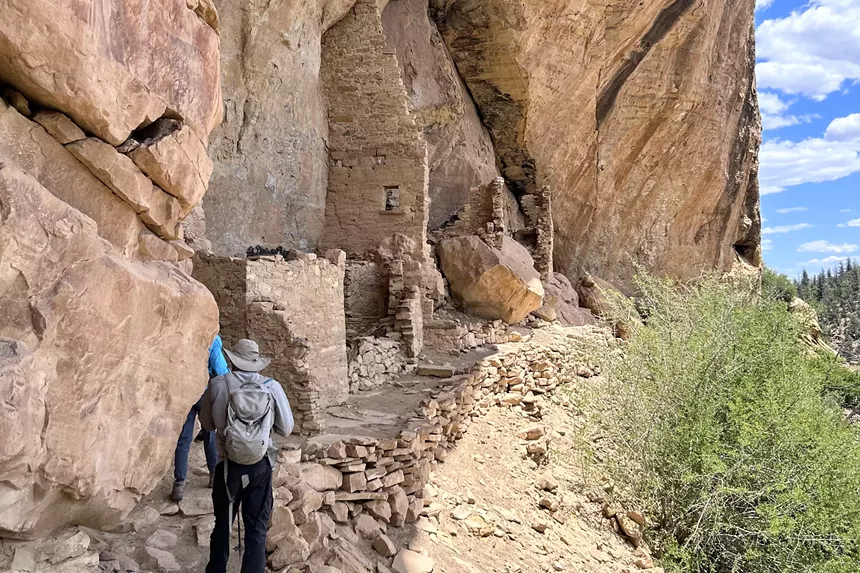 Two hikers backdropped by a cliff dwelling within Ute Mountain Tribal Park