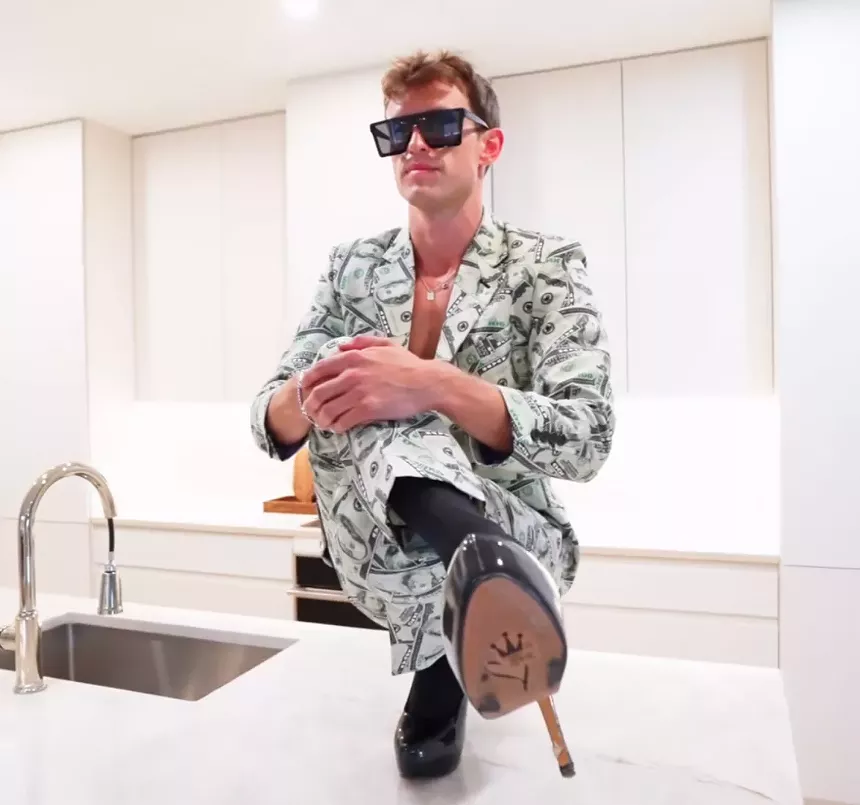 Connor Cole wear a pair of black sunglasses, an hundred-dollar bill printed suit and a pair of Patton black platform heels on a kitchen countertop.