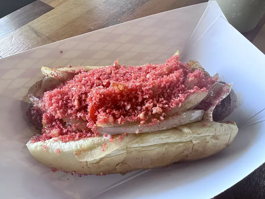 Bacon wrapped hotdog with grilled peppers and onions, topped with hot cheeto dust