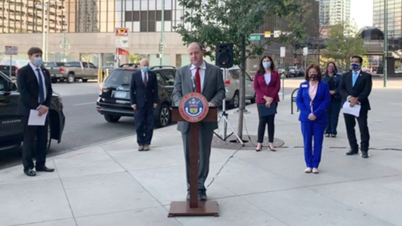 Governor Jared Polis at the podium during the October 15 press conference, as (left to right) First Gentleman Marlon Reis, Aurora Mayor Mike Coffman, Colorado Secretary of State Jena Griswold, Lieutenant Governor Dianne Primavera, State Senator Julie Gonzales and Denver Clerk and Recorder Paul López look on.