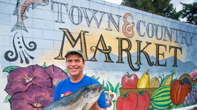 a man holds a large salmon in front of a colorful mural with fruits and vegetables