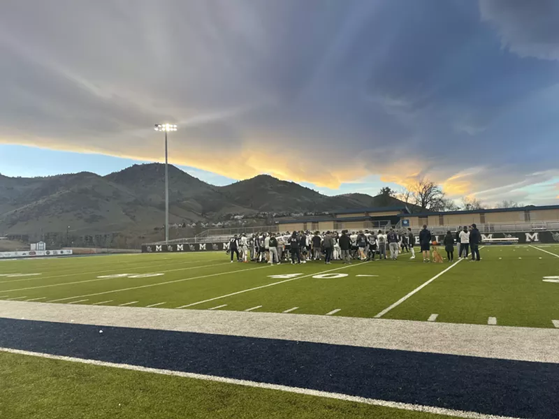 Image: Under a cloudy sky, a football team huddles on a green turf field.