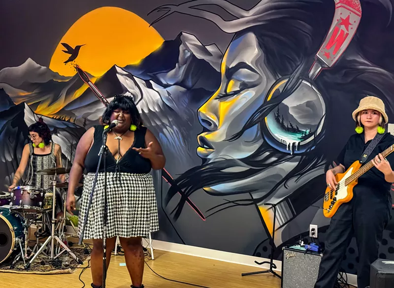 Image: a drummer, singer and bassist performing in front of a mural