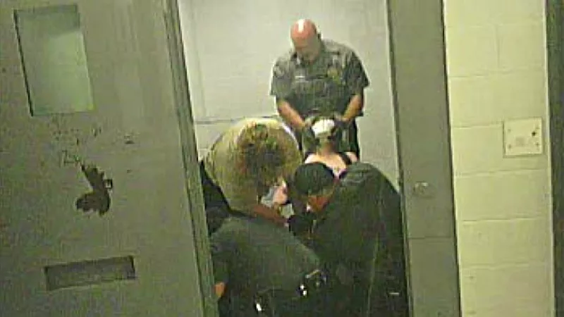 Image: In a screen capture from a video seen here, a Fremont County deputy can be seen putting a restraint hold on a naked Carolyn O'Neal, her face covered with a spit mask, as other personnel try to force her foot back into a strap on a restraint chair.