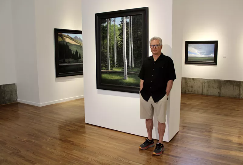 Image: man in black shirt and khaki shorts poses in a gallery with landscape paintings.