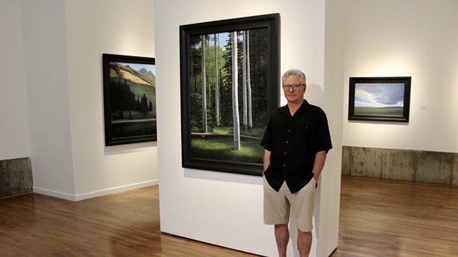 man in black shirt and khaki shorts poses in a gallery with landscape paintings.