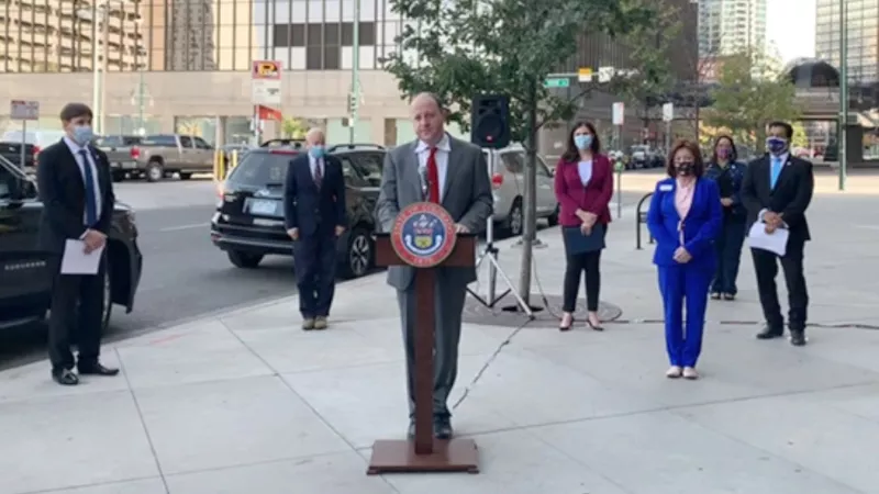 Image: Governor Jared Polis at the podium during the October 15 press conference, as (left to right) First Gentleman Marlon Reis, Aurora Mayor Mike Coffman, Colorado Secretary of State Jena Griswold, Lieutenant Governor Dianne Primavera, State Senator Julie Gonzales and Denver Clerk and Recorder Paul López look on.